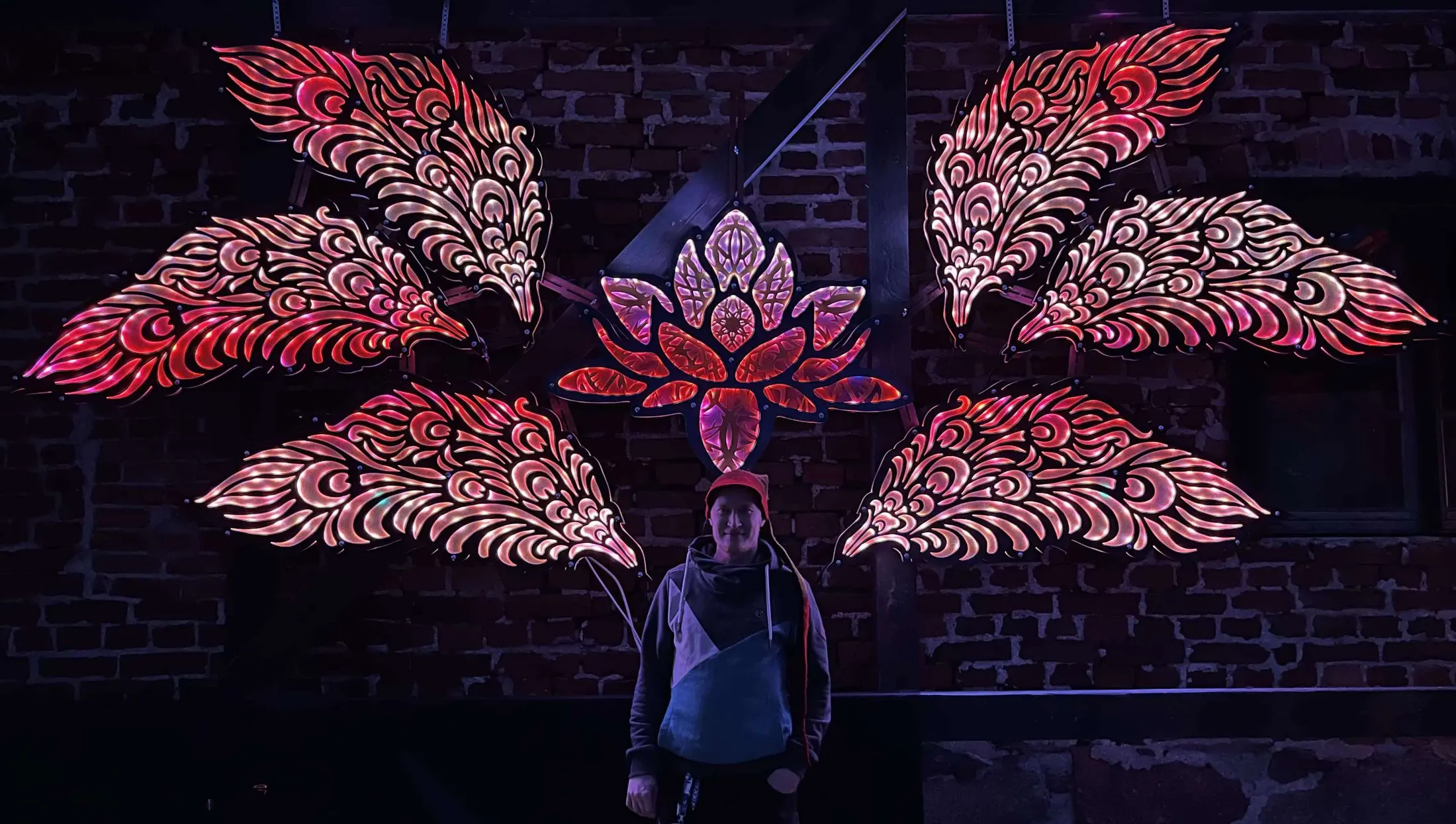 Heikki Parkkonen standing beneath an illuminated lasercut plywood sculpture of wings around a lotus, backlit with LEDs on a brick wall
