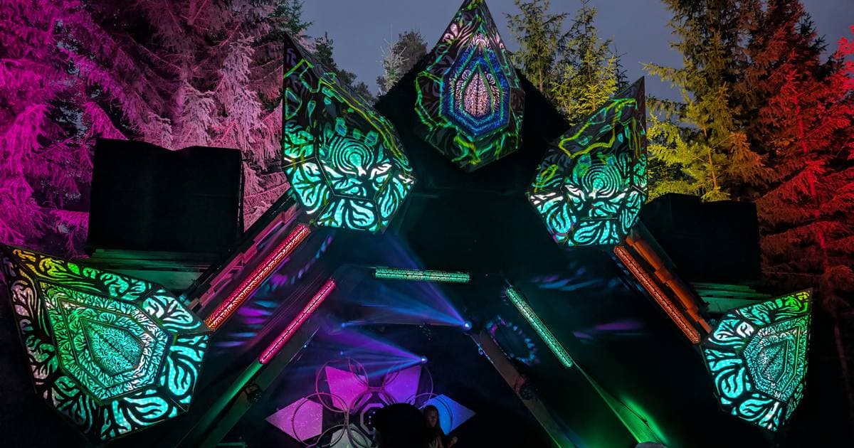 The Spacetime stage at night — five laser-cut plywood crystals lit in green and teal, the surrounding forest washed pink and red.